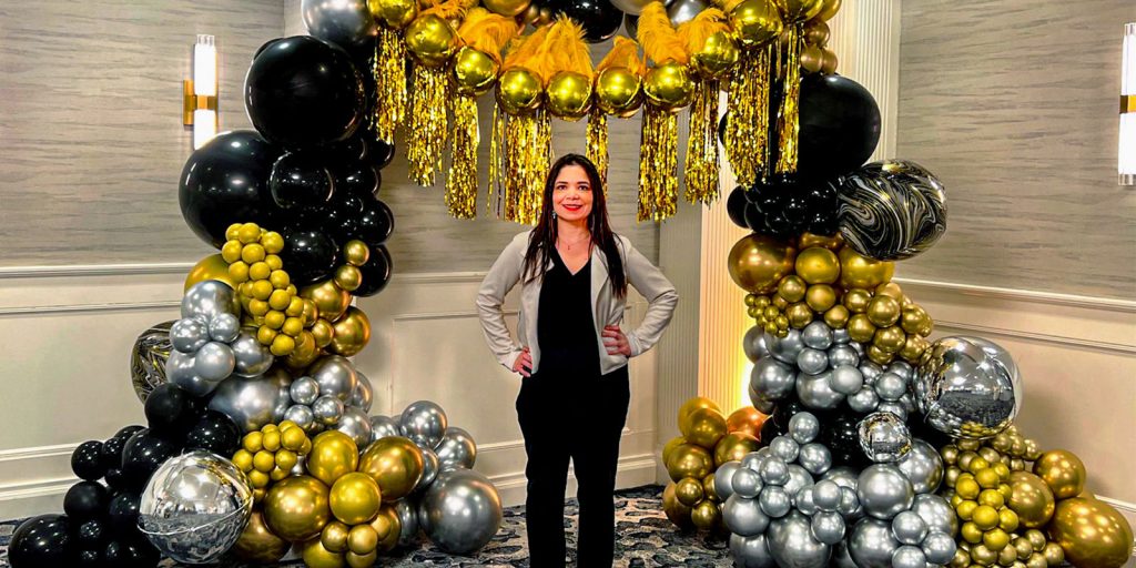 Jexsy Armstrong proudly stands before a stunning large-scale balloon arch in elegant gold and black.